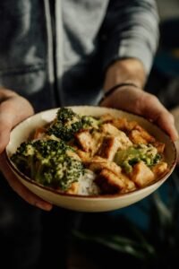 Close-up of a person holding a bowl of broccoli, tofu, and rice, offering a nutritious meal option.